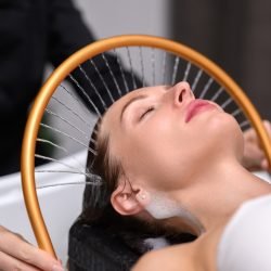 Woman undergoing a traditional Japanese head spa treatment. Gentle water streams flow through a circular device, cleansing and massaging the scalp while she relaxes with closed eyes.